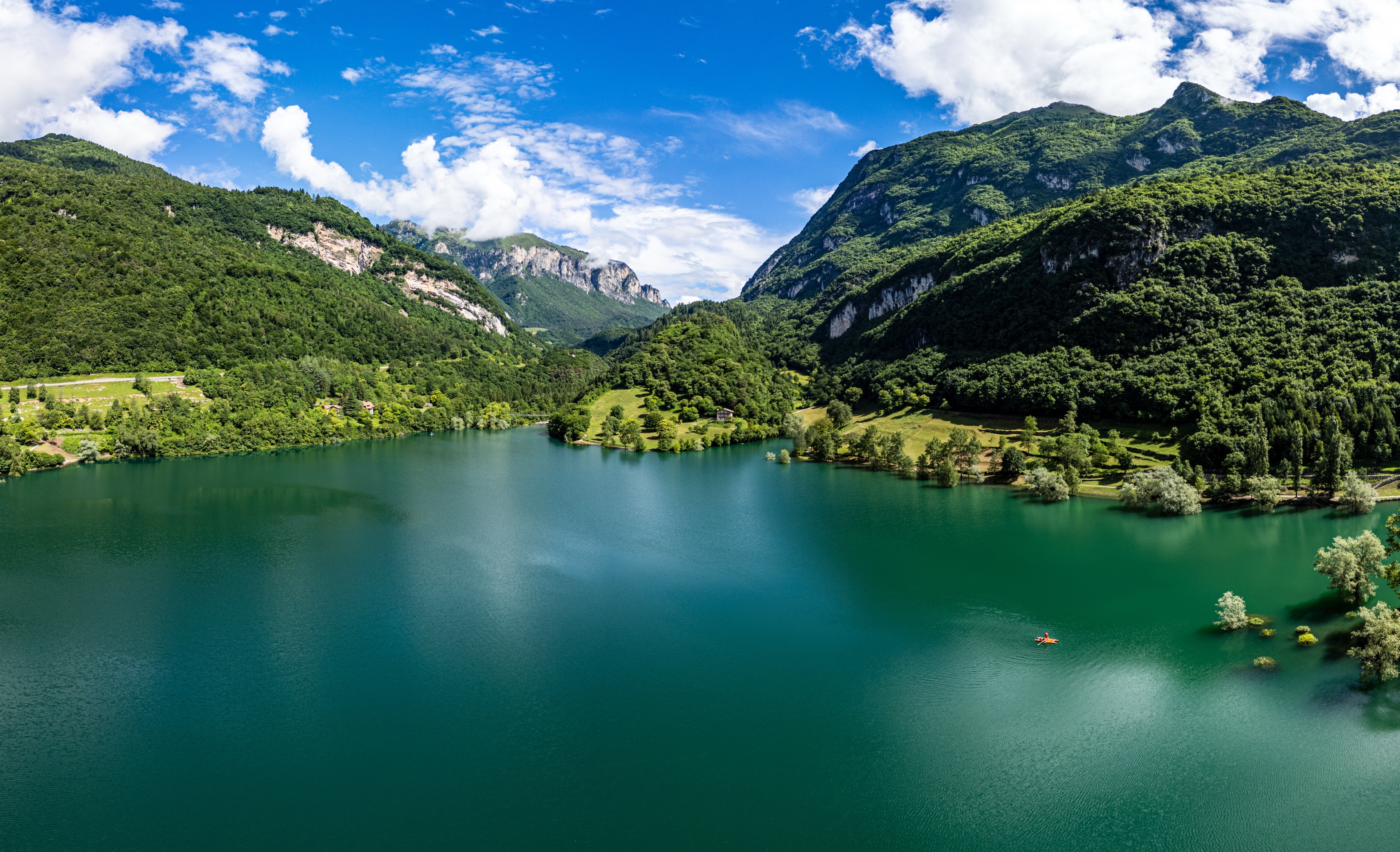 Lago di Tenno sfotografowane z drona — otoczone zielonymi wzgórzami i górami, z turkusową wodą i niewielką łódką widoczną po prawej stronie.